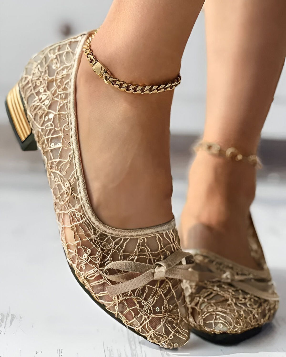 Une femme porte des chaussures plates dorées en dentelle avec un nœud sur le devant. Elle porte également une chaîne de cheville en or.