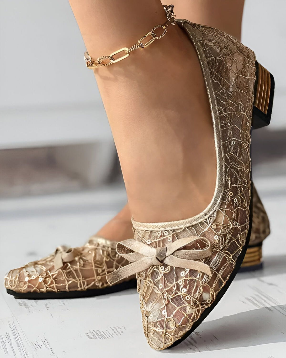Une femme porte des chaussures dorées à talons avec un nœud sur le devant. Les chaussures sont en dentelle et ont un bout pointu. La femme porte également une chaîne dorée à la cheville.