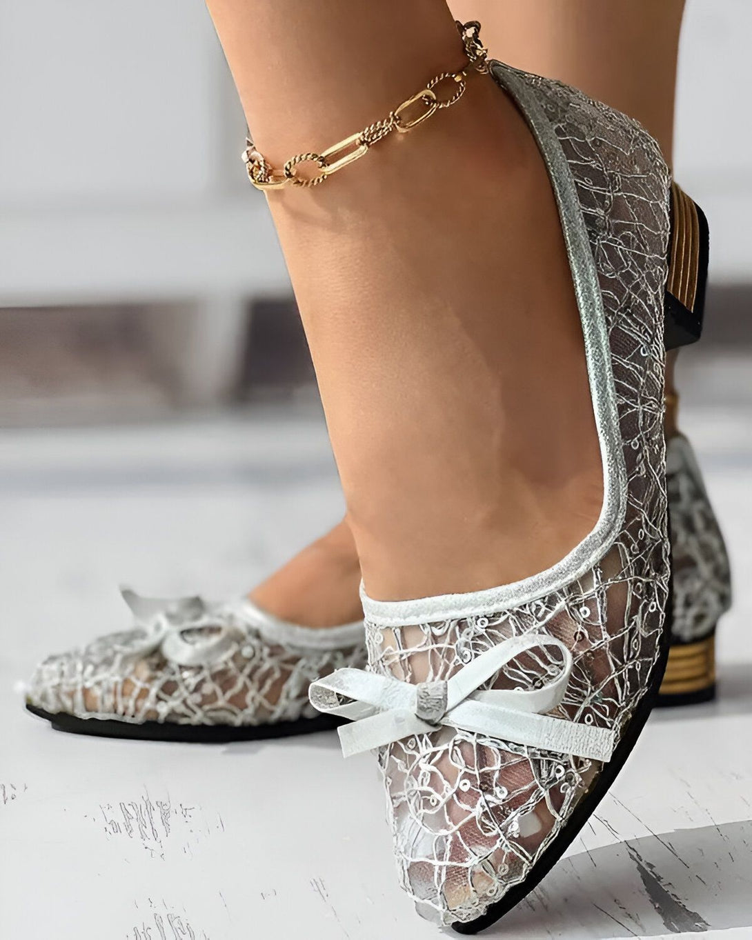 Une femme porte des chaussures plates en dentelle argentée avec un nœud blanc sur le devant. Les chaussures sont transparentes et laissent voir les orteils. La femme porte également une chaîne de cheville dorée.