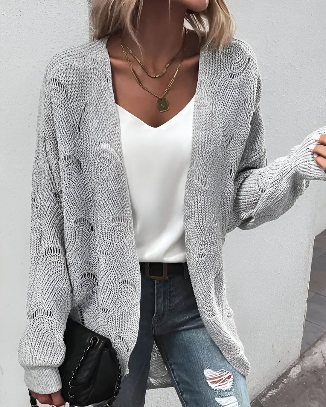 Une jeune femme porte un cardigan gris en laine, un jean bleu troué et un débardeur blanc. Elle a les cheveux blonds et porte un collier doré. Elle est debout et tient un sac à main noir.