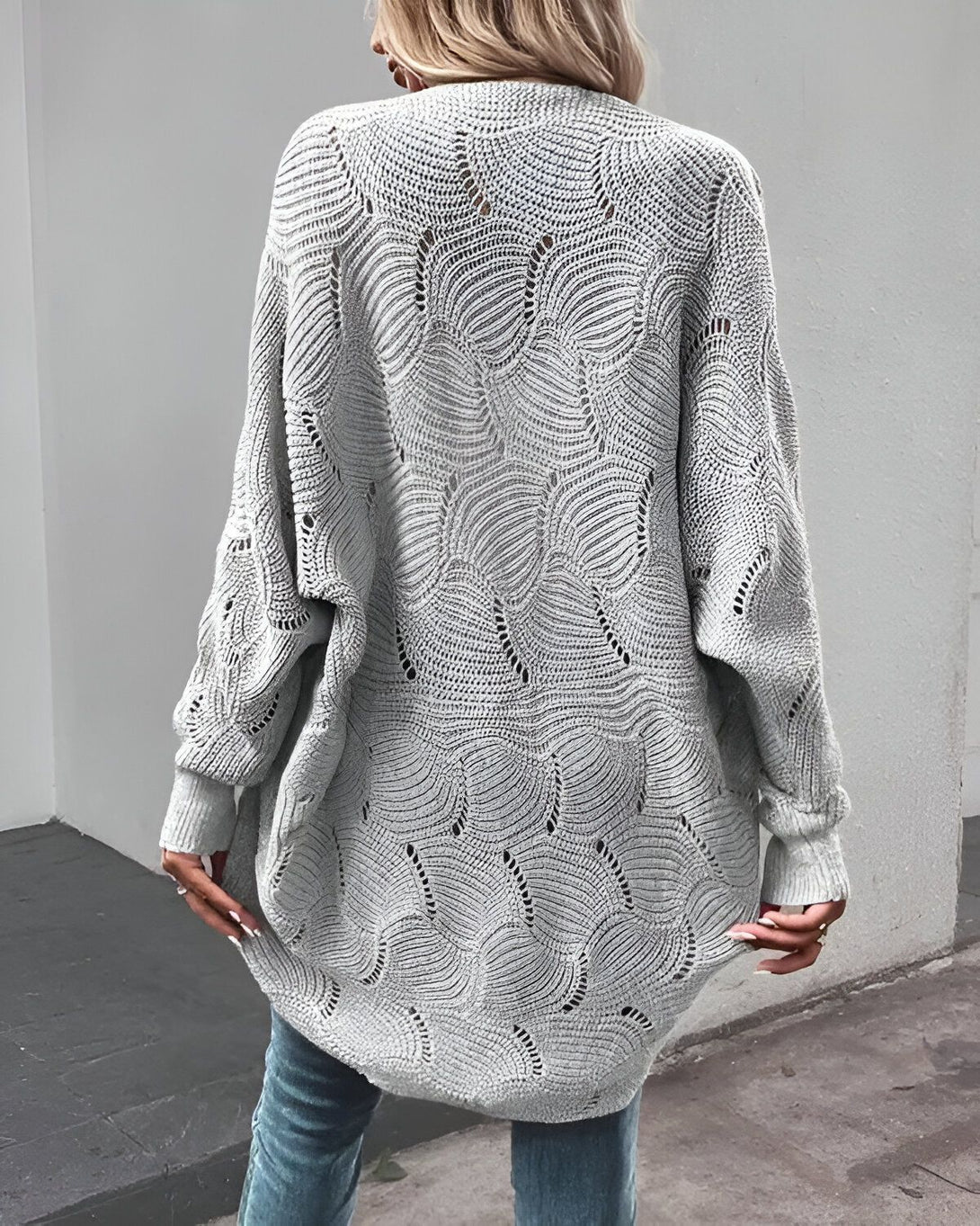 Une femme porte un cardigan gris en laine, long et ample, avec des motifs de feuilles en dentelle.