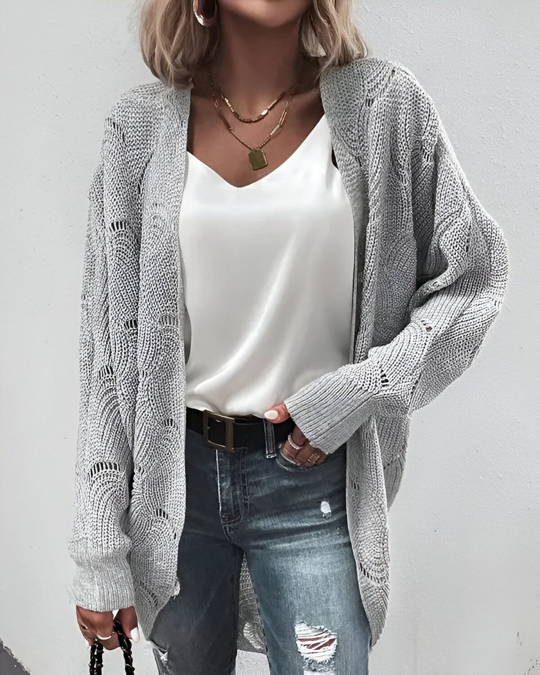 Une femme porte un gilet gris en laine, un chemisier blanc en soie, un jean bleu troué et des escarpins noirs. Elle a les cheveux blonds et un maquillage léger. Elle est debout dans une rue et sourit à la caméra.
