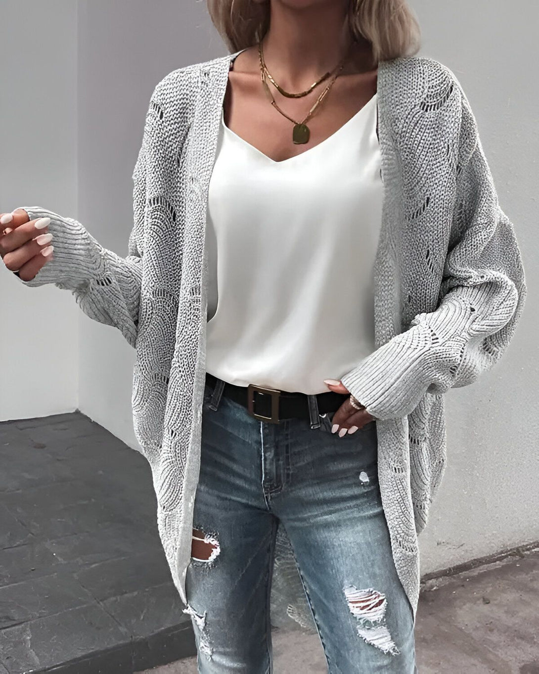 Une jeune femme porte un gilet gris en laine, un débardeur blanc en soie, un jean bleu troué et des escarpins noirs.