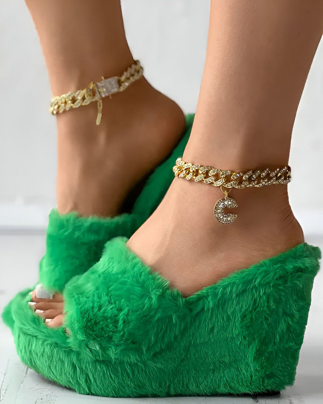 Une femme porte des mules vertes à talons compensés et à fourrure avec une chaîne de cheville en or et un pendentif en forme de C.