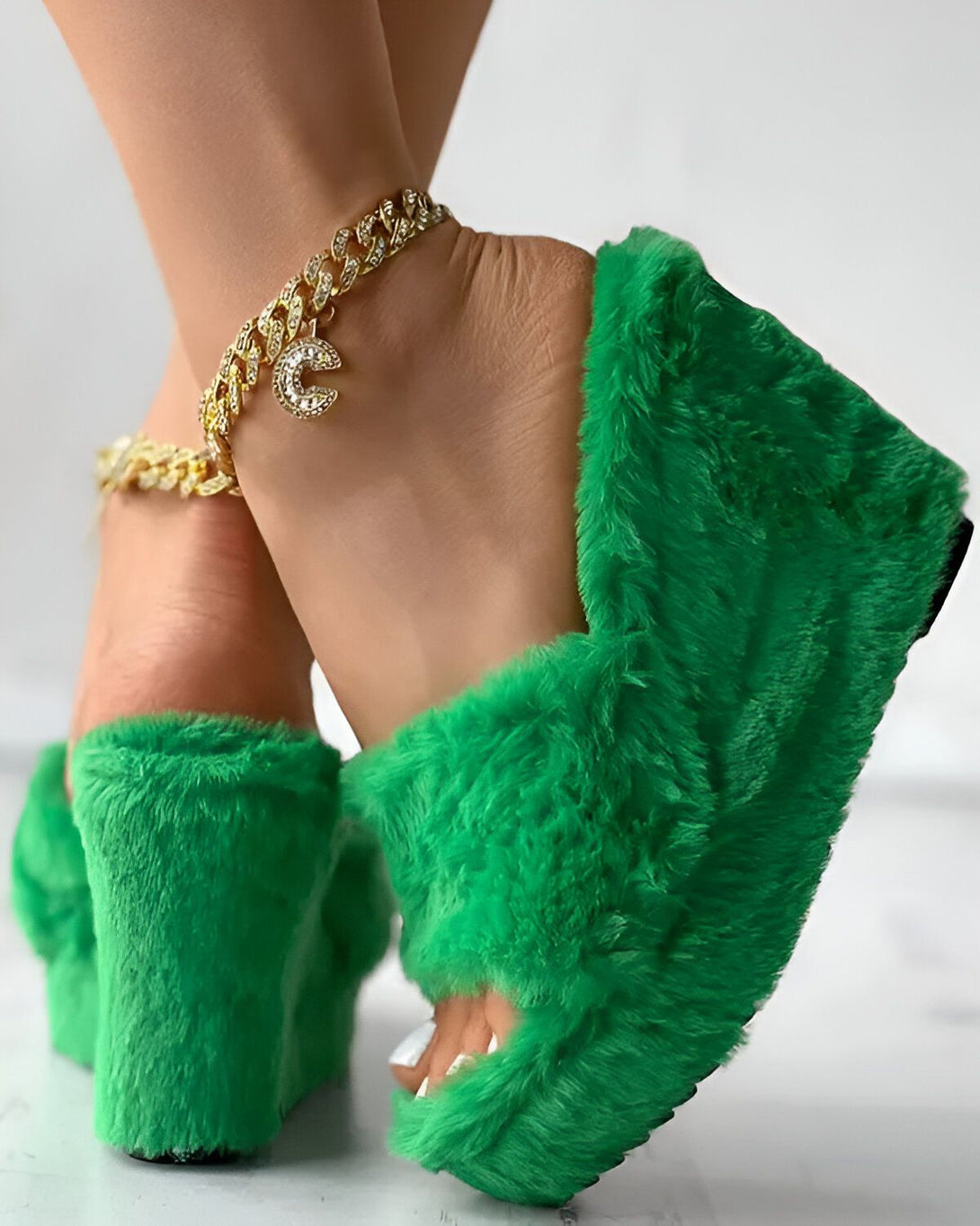 Une femme porte des sandales à talons compensés en fourrure verte. Les sandales ont une bride de cheville en chaîne dorée avec un pendentif en forme de croissant de lune. La femme a les ongles des pieds peints en vert.