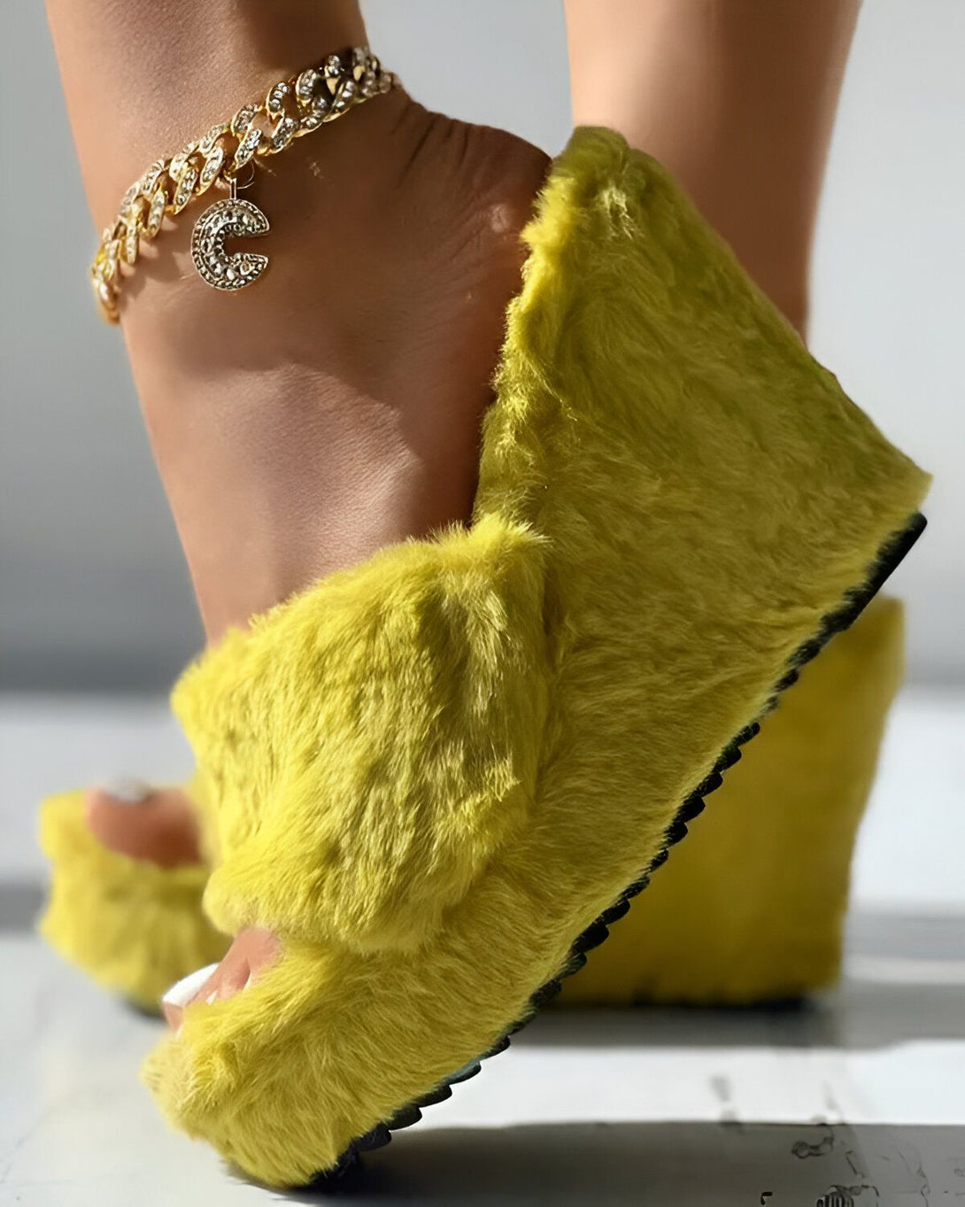 Une femme porte des sandales à talons compensés jaunes. Les sandales sont en fourrure et ont une bride à boucle. La femme porte également une cheville en or avec un pendentif en forme de C.