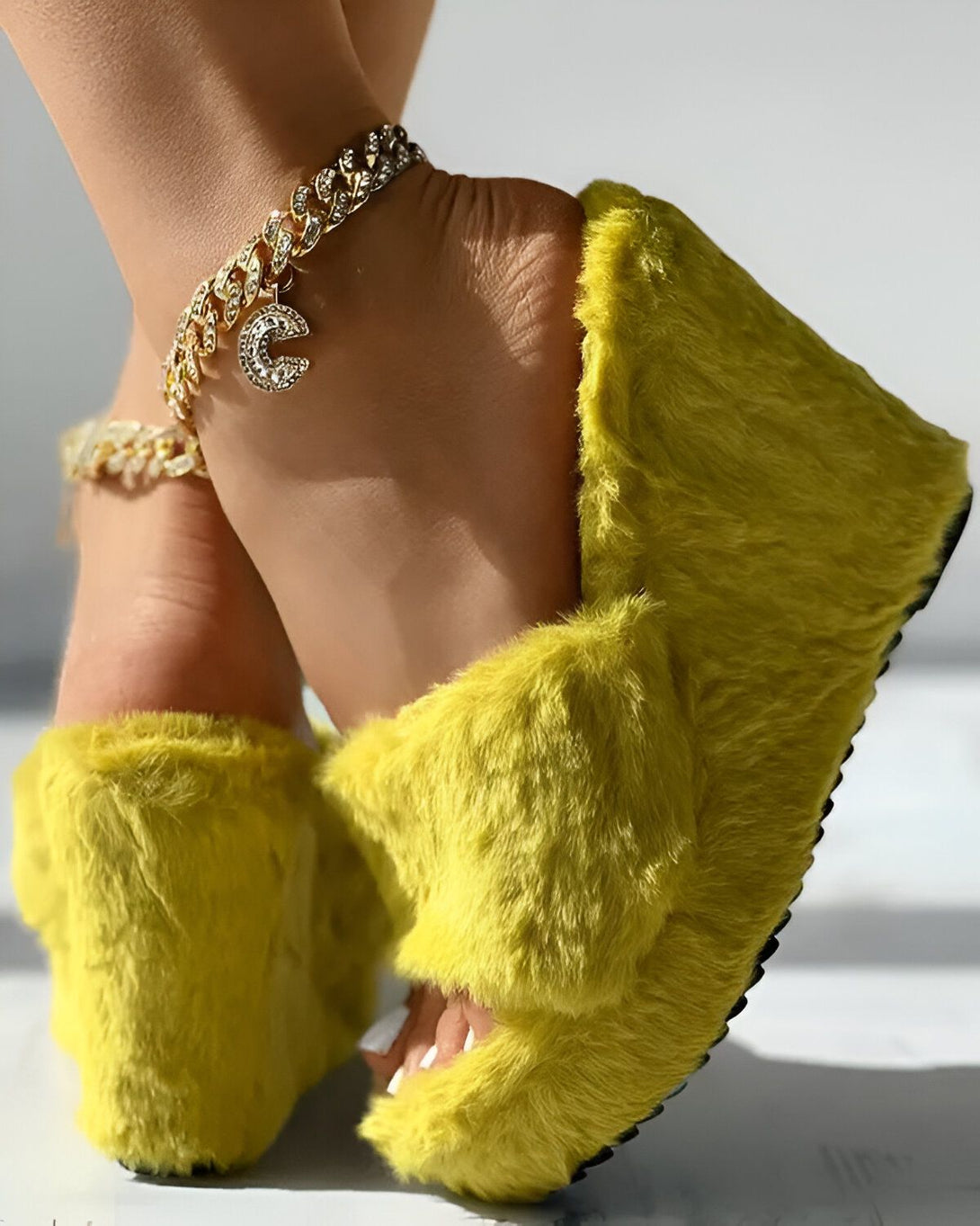 Une femme porte des sandales jaunes à talons compensés en fourrure. Elle porte également un bracelet de cheville en or avec un pendentif en forme de C.
