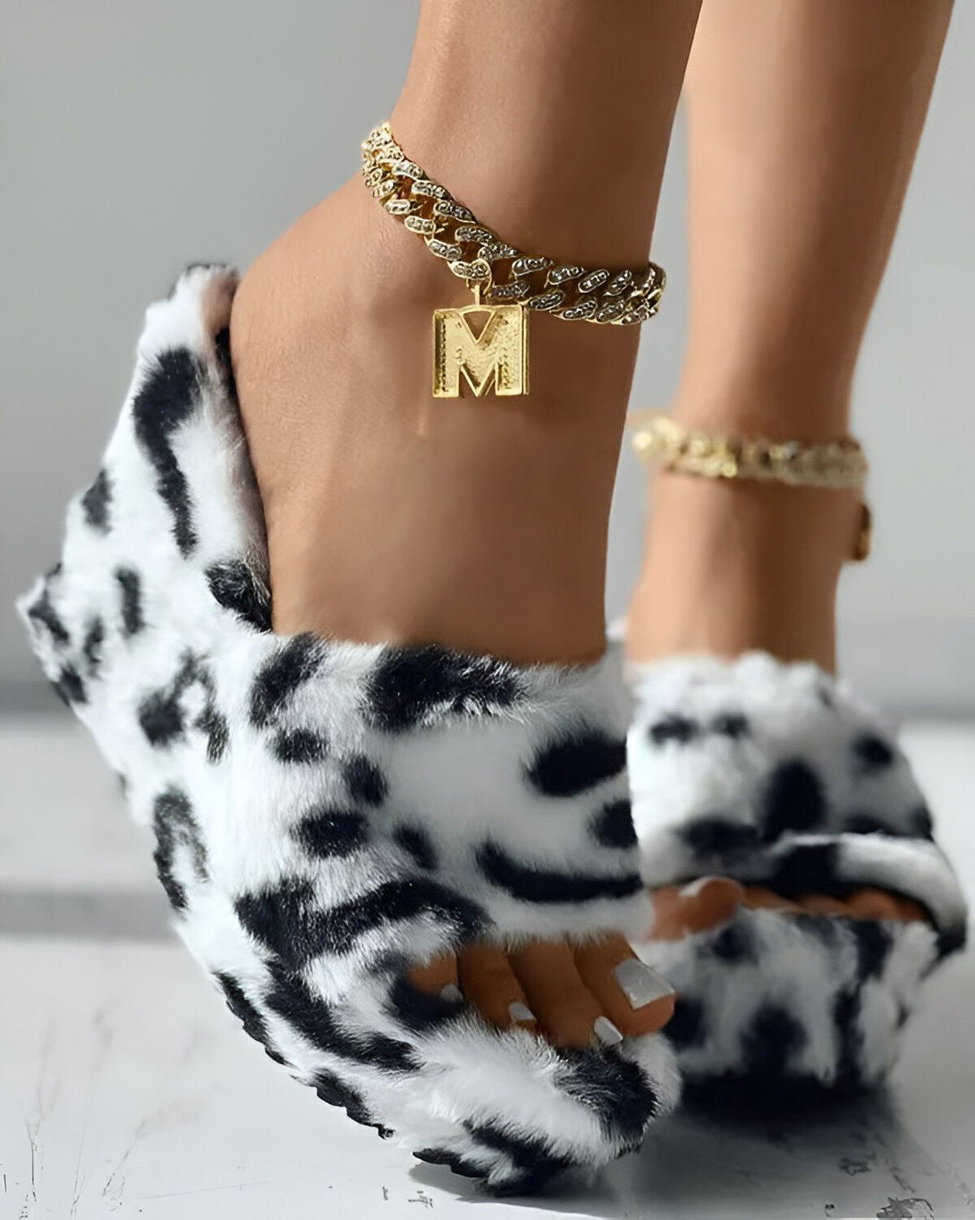 Une image d'une paire de mules à imprimé vache en fausse fourrure blanche et noire. Une chaîne de cheville en or avec un pendentif en forme de M est portée sur le pied droit.