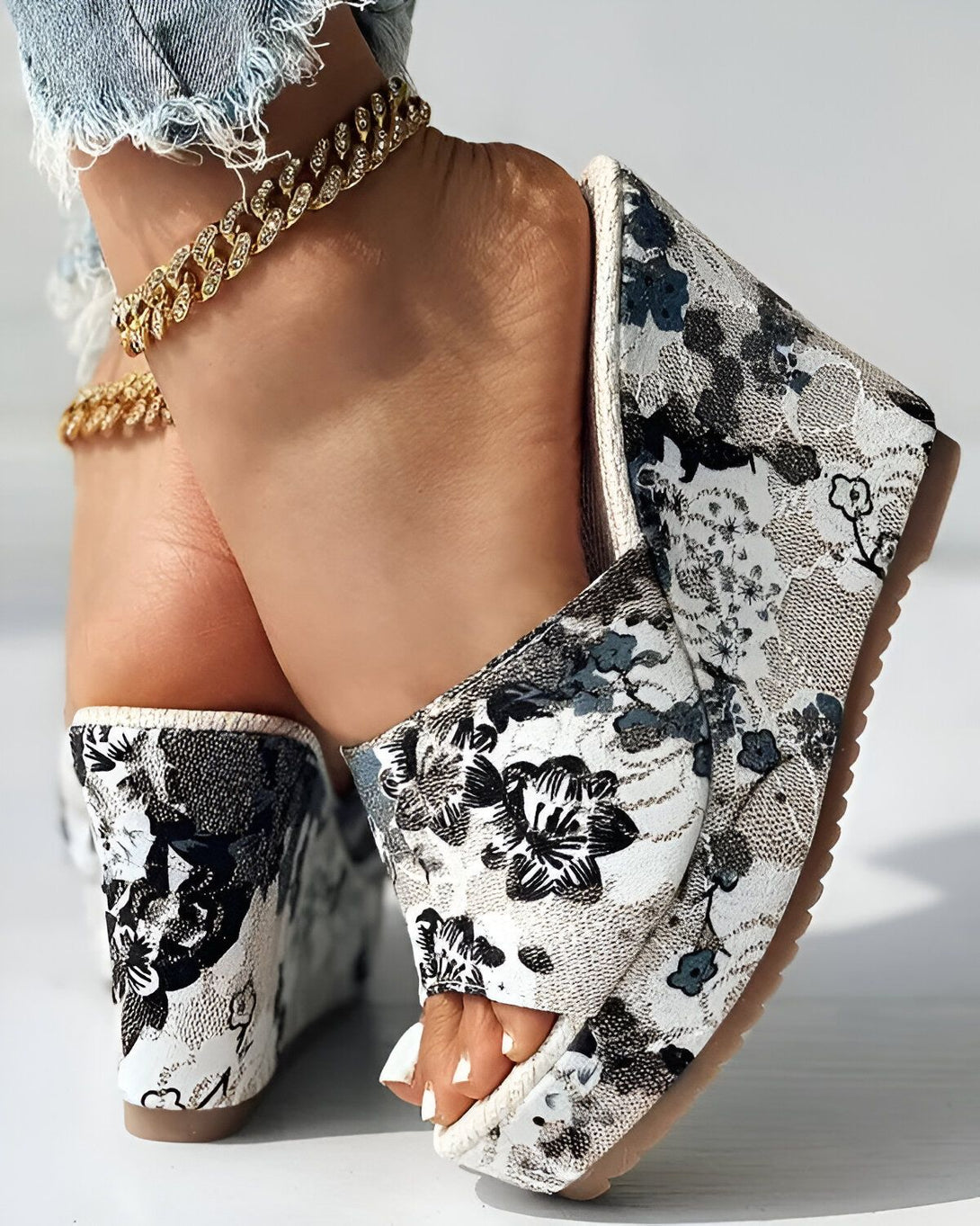 Une femme porte des mules à talons compensés à motif floral noir et blanc. Elle porte également un jean bleu déchiré et un bracelet de cheville en or.
