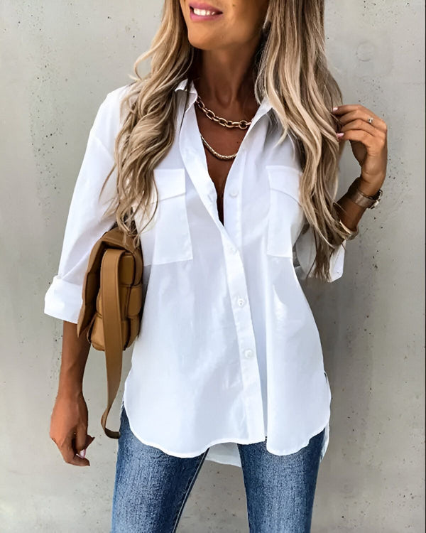 Une jeune femme porte une chemise blanche ample et des jeans bleus. Elle a les cheveux longs et blonds et porte un collier doré. Elle porte également un sac à main marron.