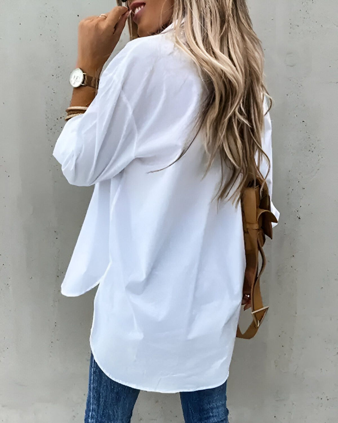 Une femme porte une chemise blanche surdimensionnée avec des manches retroussées et un jean bleu. Elle porte également un sac à dos marron et une montre en métal argenté. Ses cheveux sont longs et blonds et elle porte un maquillage naturel.