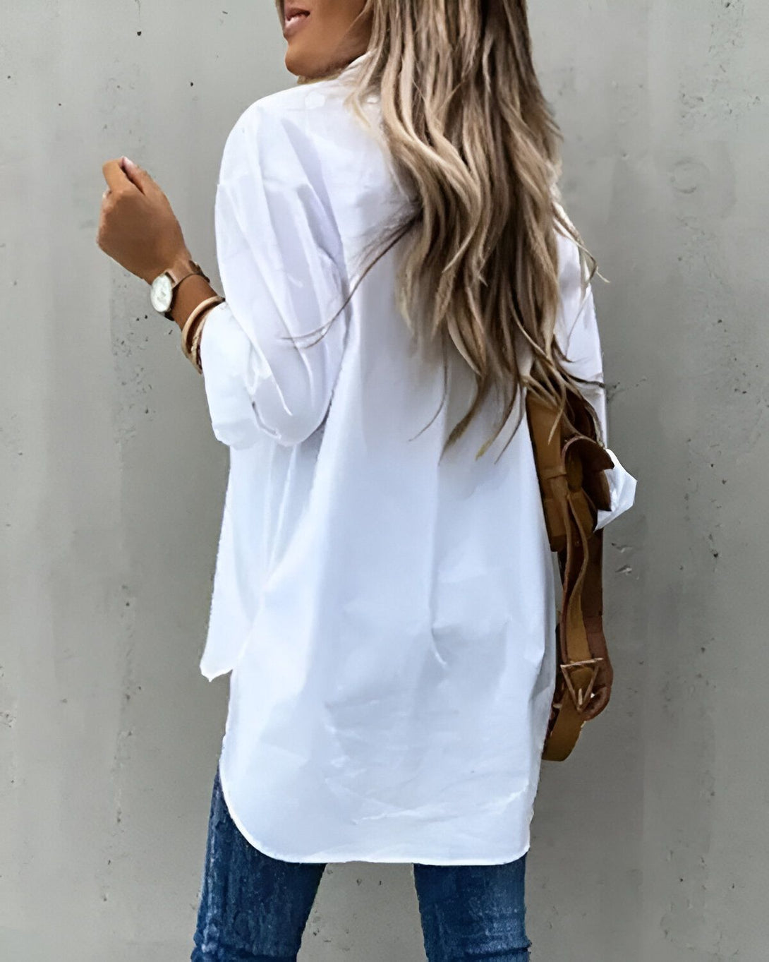 Une femme porte une chemise blanche oversize avec un jean bleu et un sac à dos marron. Elle a les cheveux longs et blonds et porte une montre au poignet gauche.