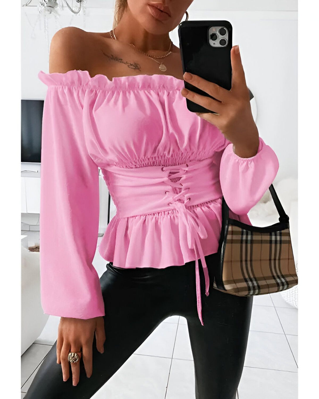 Une jeune femme porte un chemisier rose à épaules dénudées. Le chemisier est froncé sur le devant et possède des manches longues. Elle porte également un pantalon noir et un sac à main marron.