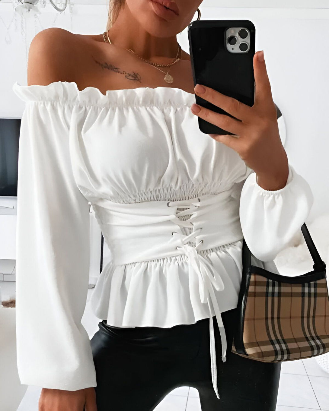 Une jeune femme porte un chemisier blanc à épaules dénudées et à lacets dans le dos. Elle porte également un pantalon noir et un sac à main marron.