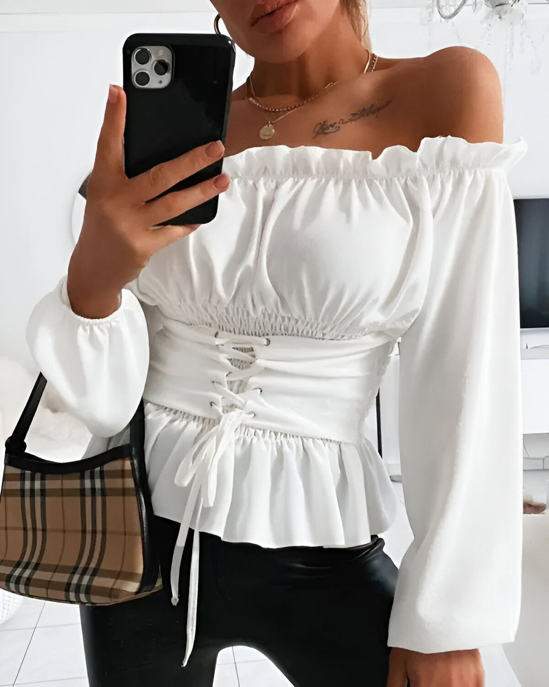 Une jeune femme porte un chemisier blanc à épaules dénudées et à lacets dans le dos. Elle porte également un pantalon noir et un sac à main marron.