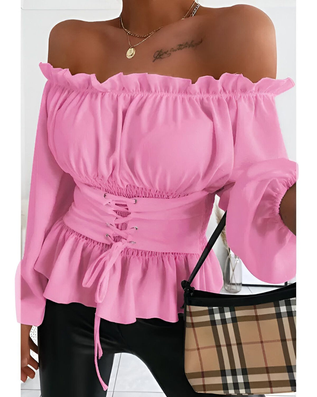 Une femme porte un chemisier rose à épaules dénudées. Le chemisier est froncé à la taille et possède des manches bouffantes. Elle porte également un pantalon noir et un sac à main marron.