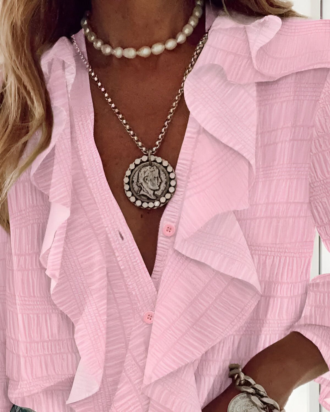 Une femme porte une chemise rose à volants avec un collier de perles et un pendentif rond en métal.