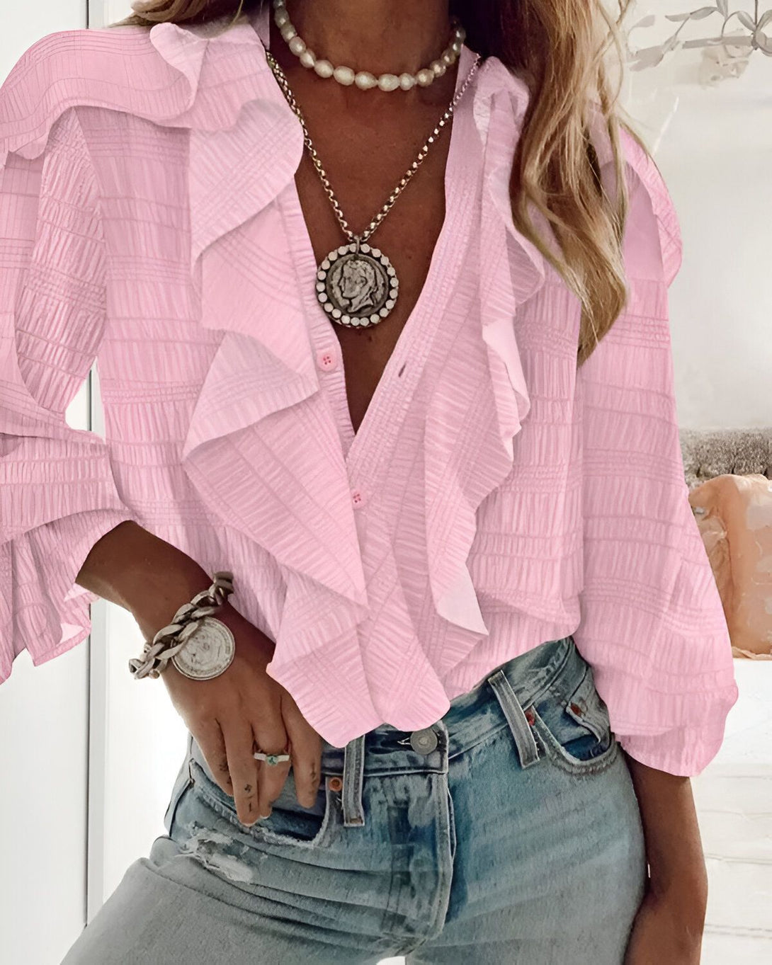 Une femme porte une chemise rose à volants avec un jean bleu et un collier de perles.