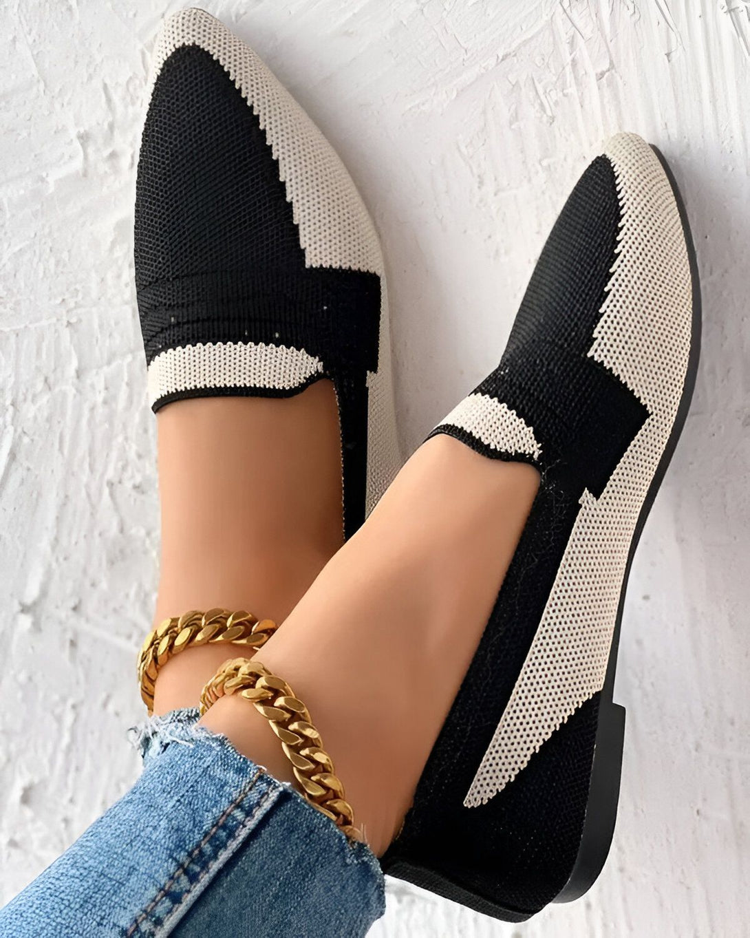 Une image de chaussures plates noires et blanches avec un motif géométrique. Les chaussures sont portées par une femme qui porte un jean bleu et un bracelet de cheville doré. Le fond est blanc.