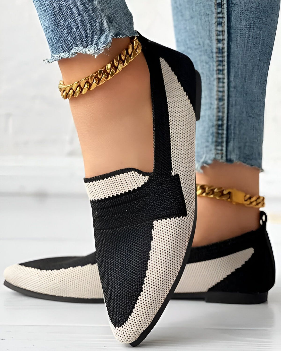 Une femme porte des chaussures plates noires et blanches avec un jean bleu et des chaussettes blanches. Elle porte également deux bracelets de cheville en or.