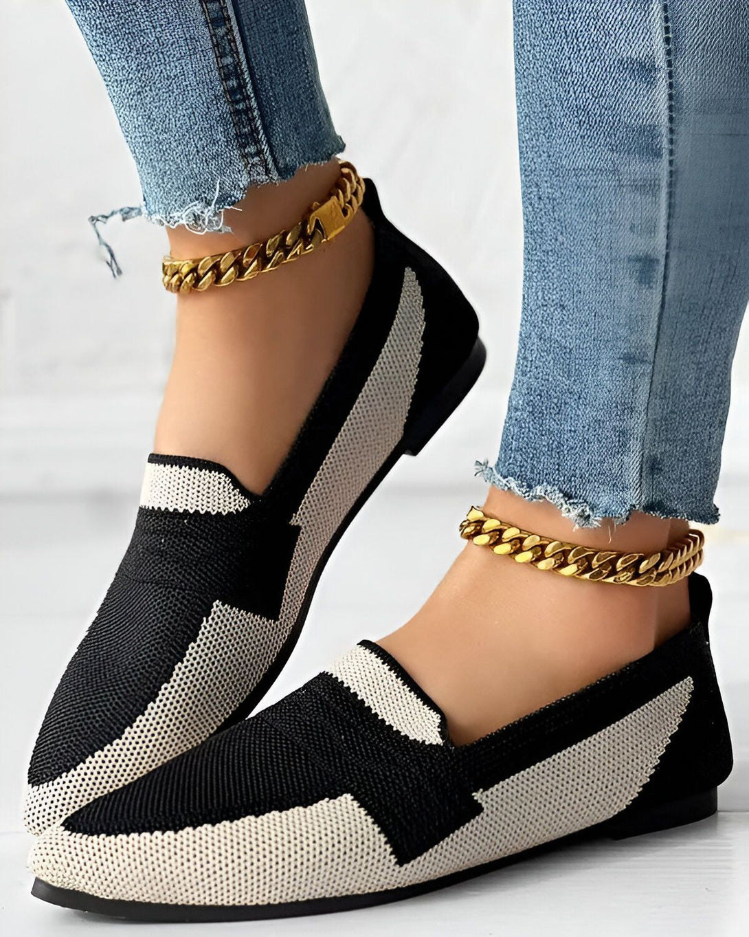 Une femme porte des chaussures plates noires et blanches avec un jean bleu et des bracelets de cheville en or.