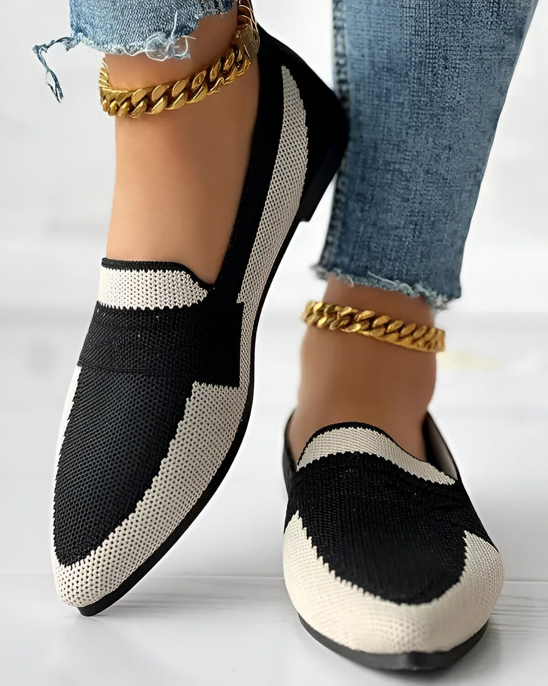 Une femme porte des chaussures plates noires et blanches avec un jean bleu et des chaussettes noires. Elle porte également une chaîne de cheville en or.
