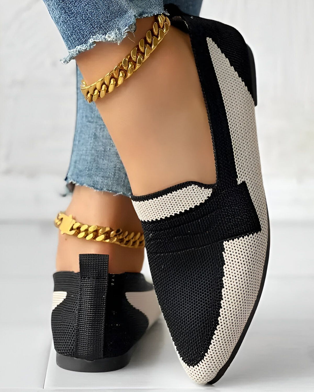 Une femme porte des chaussures plates noires et blanches avec un motif géométrique. Elle porte également un jean bleu et un bracelet de cheville doré.