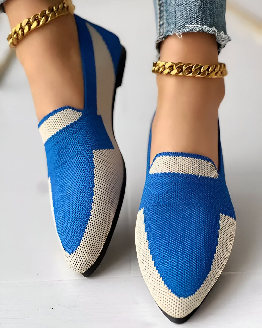 Une image de chaussures bleues et blanches avec un bout pointu et une semelle plate. Les chaussures sont portées par une femme qui porte un jean et des chaussettes blanches. La femme est debout sur un sol en béton.