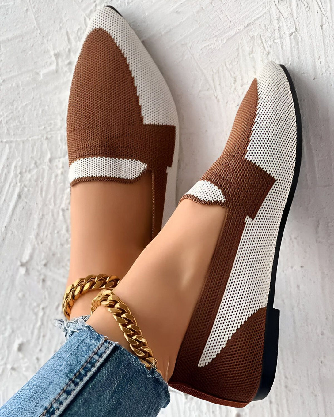Une image de chaussures plates marron et blanches avec un motif en maille. Les chaussures sont portées par une femme qui porte des jeans et un bracelet de cheville en or. Le fond est un mur blanc.