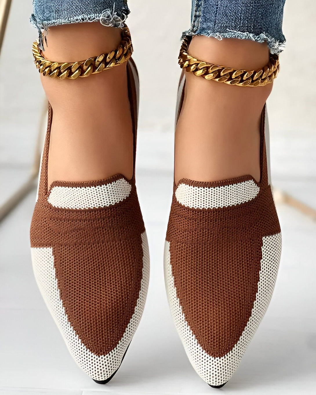 Une femme porte des chaussures marron et blanches avec un bout pointu et un talon plat. Elle porte également des chaussettes blanches et un bracelet de cheville doré.