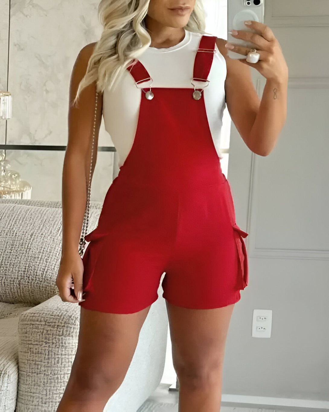 Une femme porte une combinaison-short rouge. La combinaison-short est dotée de bretelles réglables, de poches cargo et d'une fermeture à glissière sur le devant. La femme est blonde et porte un débardeur blanc. Elle est debout dans un salon et se prend en photo dans un miroir.