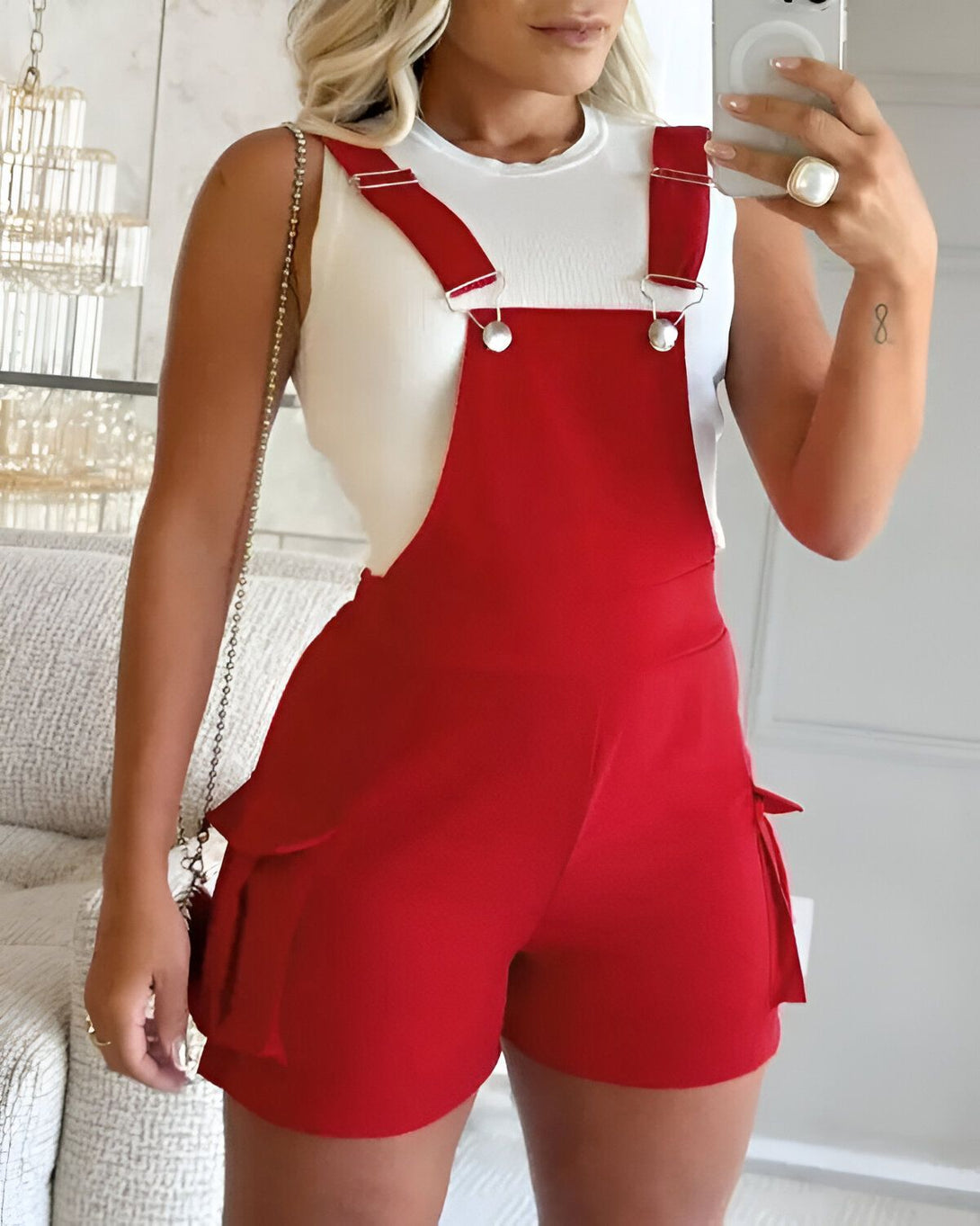 Une femme porte une combinaison-short rouge. Elle est de couleur rouge et possède des poches sur les côtés. La femme est blonde et porte un débardeur blanc. Elle est debout dans un salon et se prend en photo avec son téléphone.