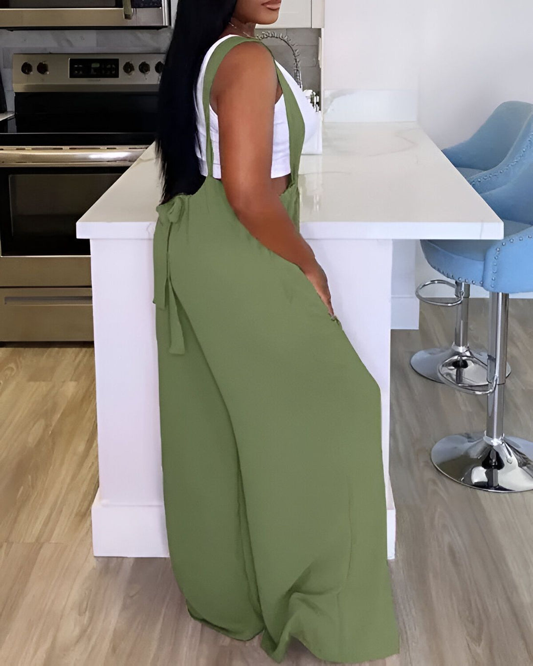 Une femme porte une combinaison-pantalon vert olive avec un haut blanc. Elle est vue de dos et est debout dans une cuisine.
