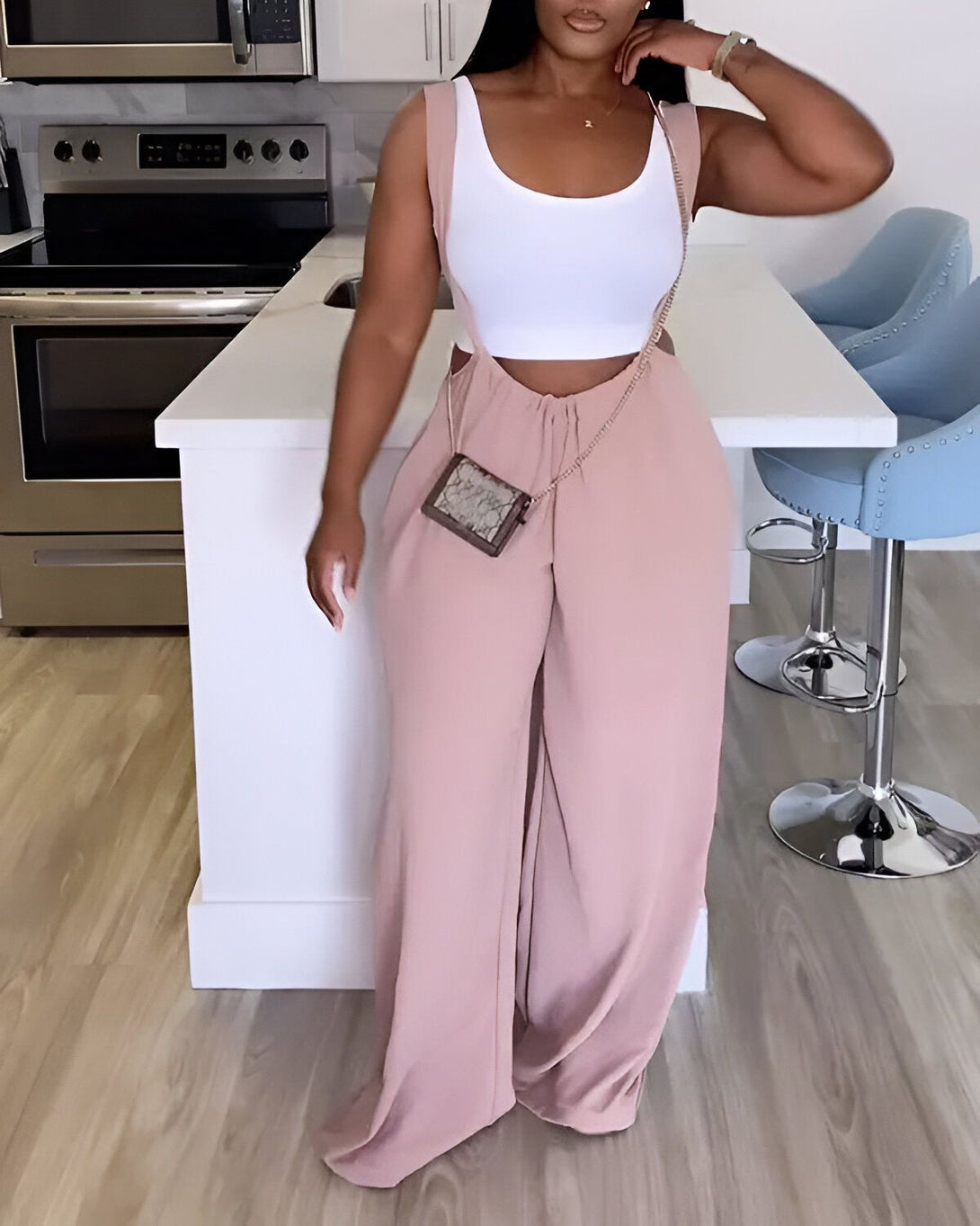 Une femme est debout dans une cuisine. Elle porte un pantalon rose taille haute et un débardeur blanc. Elle porte également un sac à main marron.