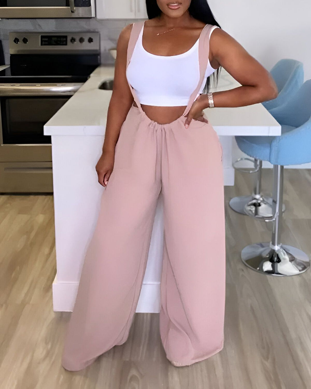 Une femme porte un pantalon large rose et un débardeur blanc. Elle est debout dans une cuisine.
