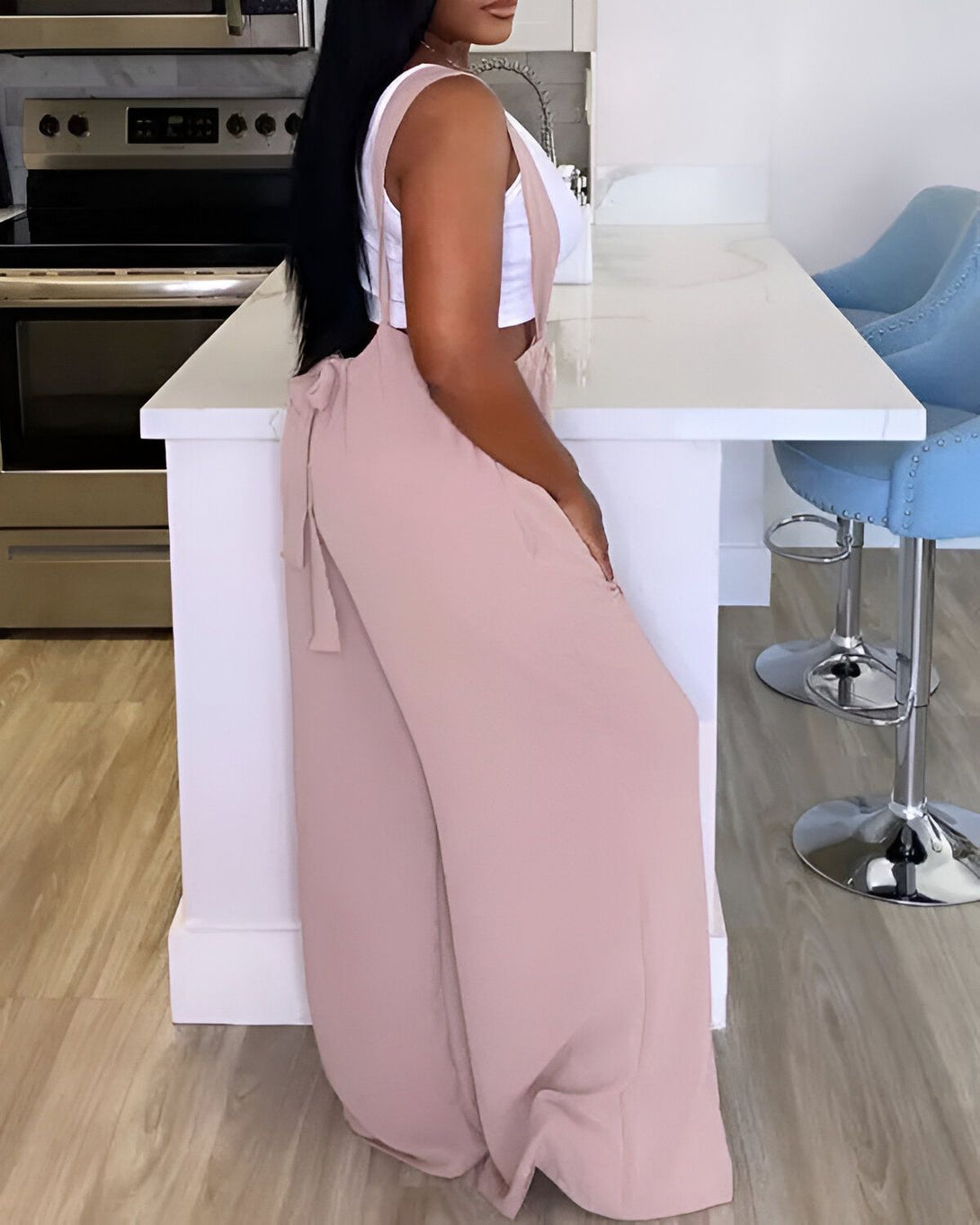 Une femme est debout dans une cuisine. Elle porte une combinaison pantalon beige et un débardeur blanc. Elle est vue de côté.
