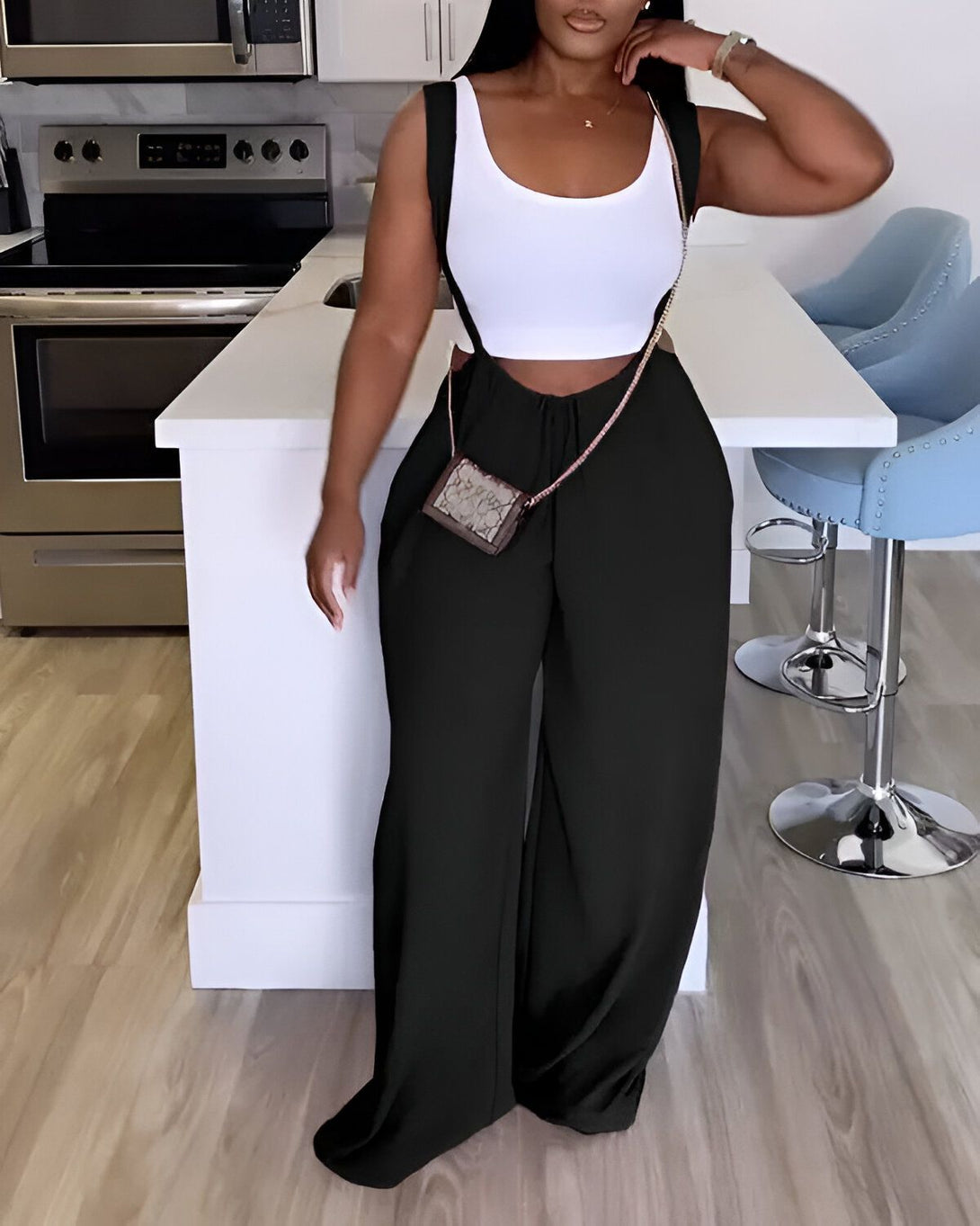 Une femme porte un pantalon noir et un haut blanc. Elle est debout dans une cuisine et porte un sac à main marron.