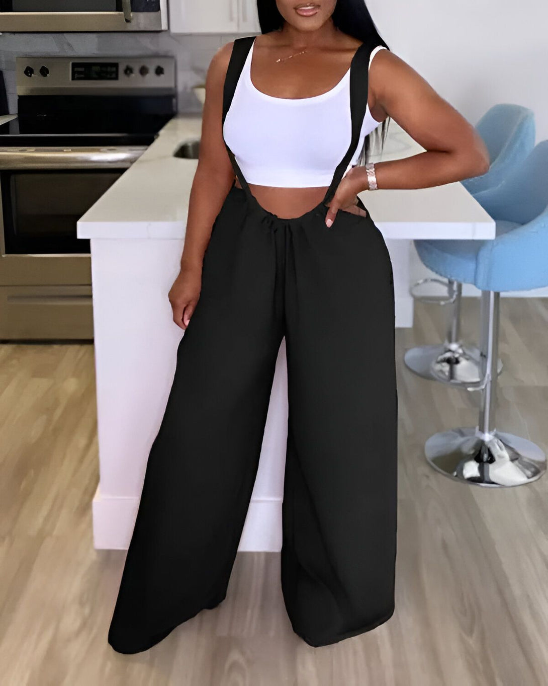 Une femme porte une combinaison pantalon noire avec un haut blanc. Elle est debout dans une cuisine.