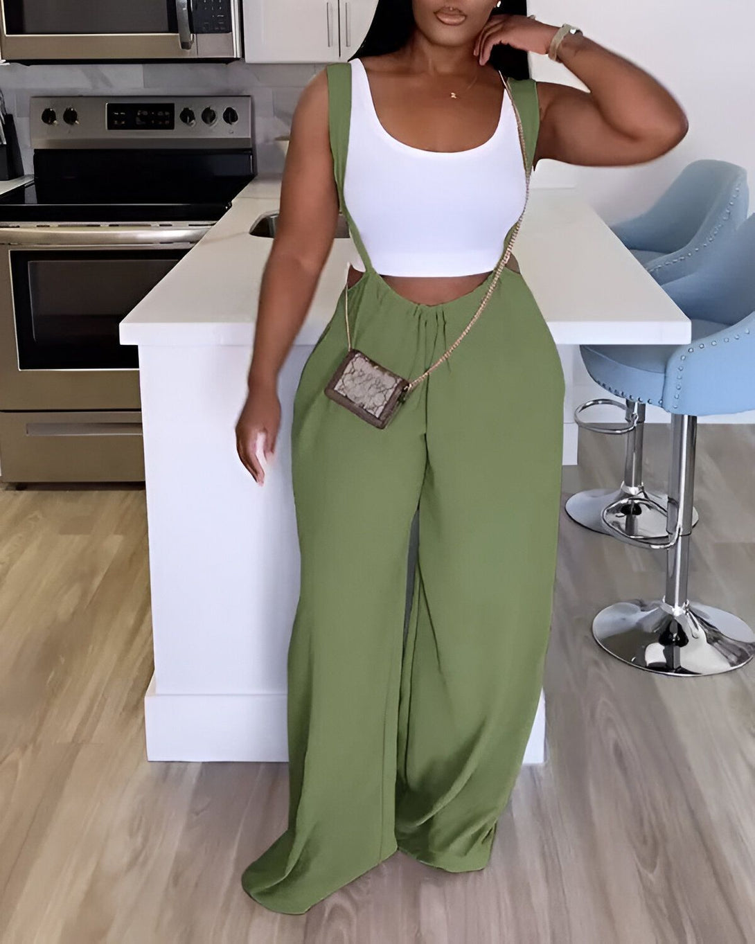 Une femme porte un pantalon vert olive taille haute avec une chaîne à la taille et un haut blanc court. Elle porte également un sac à main marron et des talons hauts.