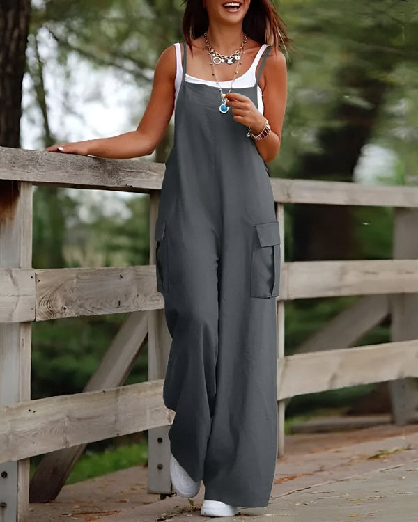 Une femme porte une combinaison-pantalon gris avec des poches et des bretelles. Elle est pieds nus et porte un collier. Elle est debout sur un pont en bois et sourit.