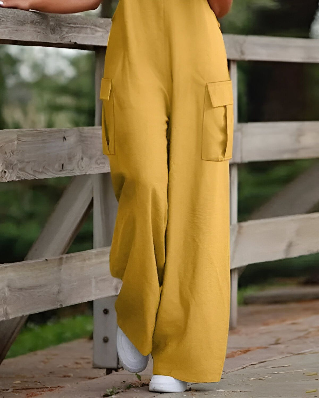 Une femme porte une combinaison-pantalon jaune moutarde ample et des baskets blanches. Elle est debout sur un pont en bois et tient le garde-corps.