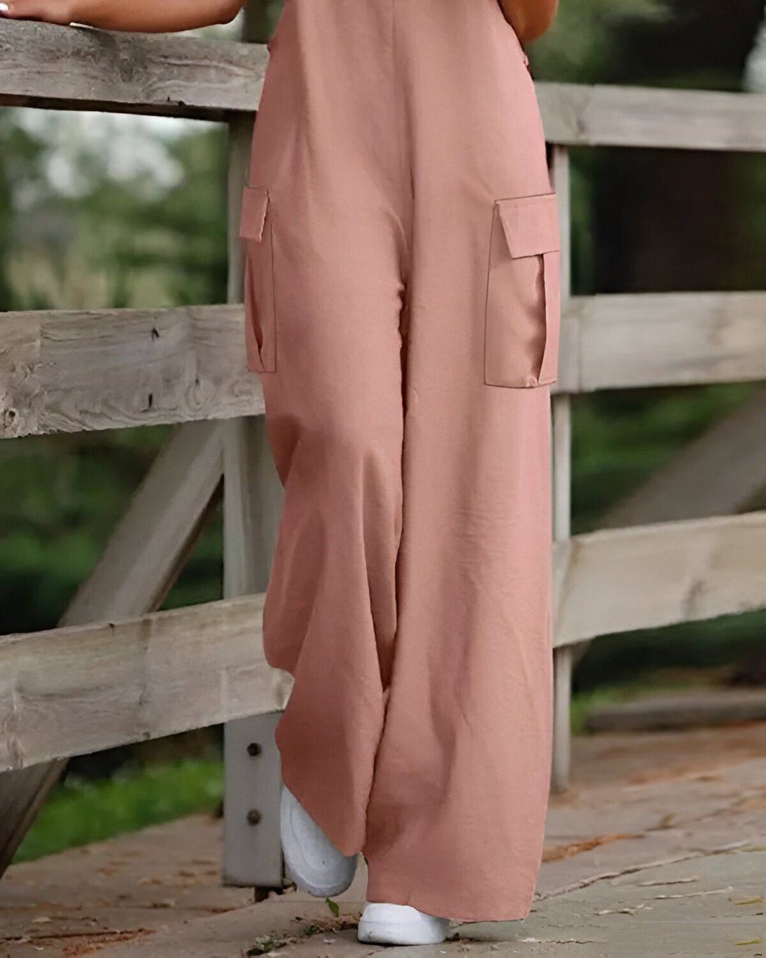 Une femme porte une combinaison-pantalon rose avec des poches et des baskets blanches. Elle est debout sur un pont en bois et tient le garde-corps.