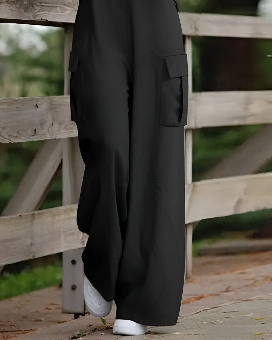 Une image d'une femme portant un pantalon cargo noir ample avec des poches sur les côtés. Elle est debout sur un pont en bois avec des arbres en arrière-plan.