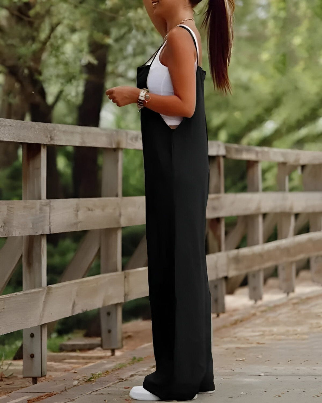 Une femme portant une combinaison noire est debout sur un pont en bois. Elle a les cheveux longs et bruns et porte un sac à main noir.
