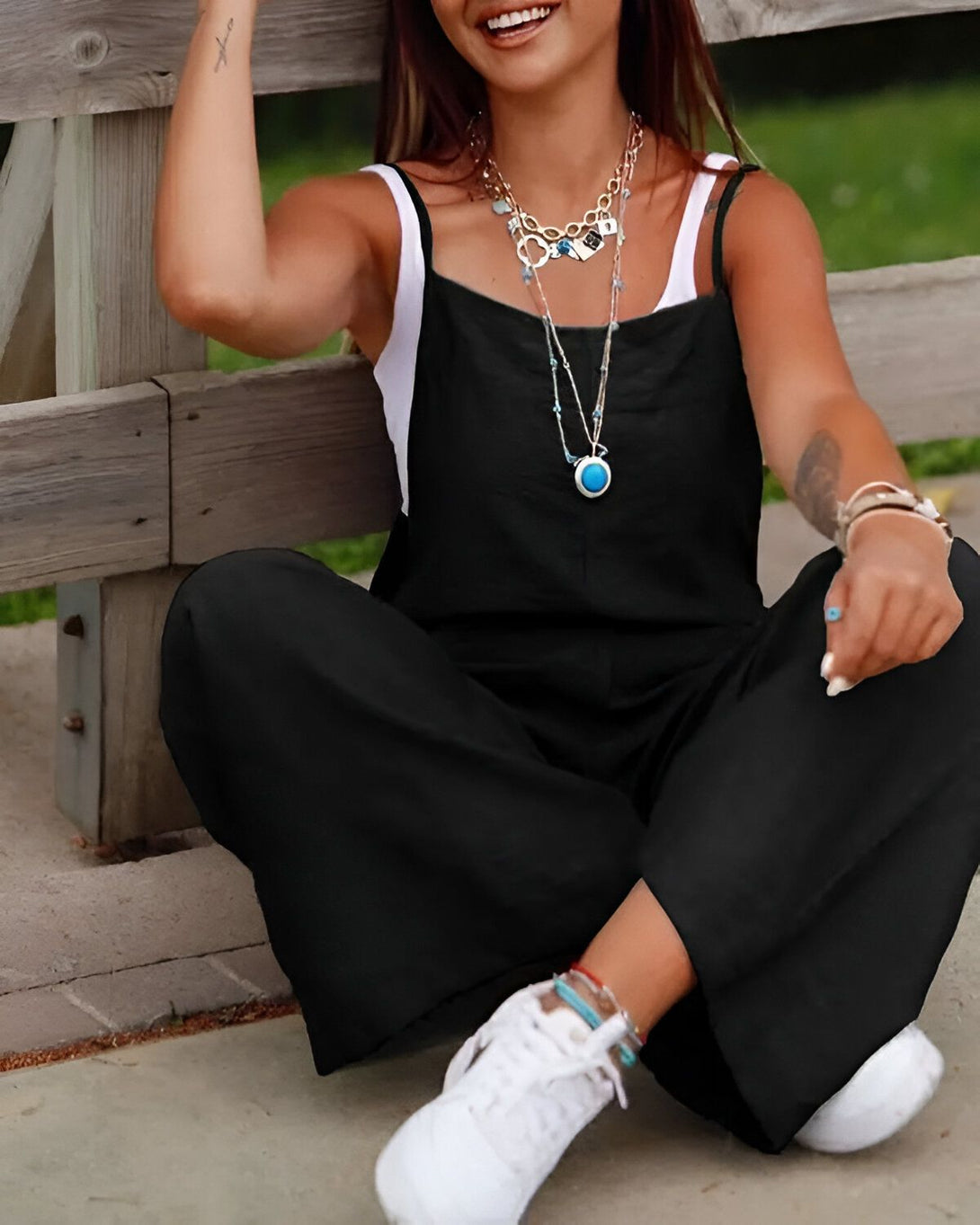 Une jeune femme est assise sur un banc en bois. Elle porte une combinaison noire, un collier avec un pendentif bleu et des baskets blanches. Elle a les cheveux longs et bruns et elle sourit.