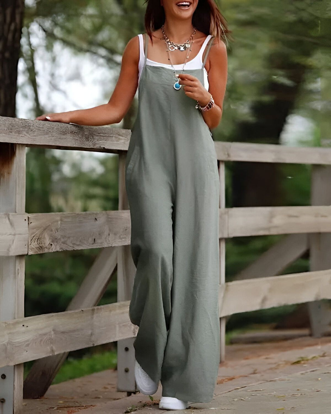 Une femme porte une combinaison-pantalon vert kaki avec un débardeur blanc. Elle est debout sur un pont en bois et sourit.