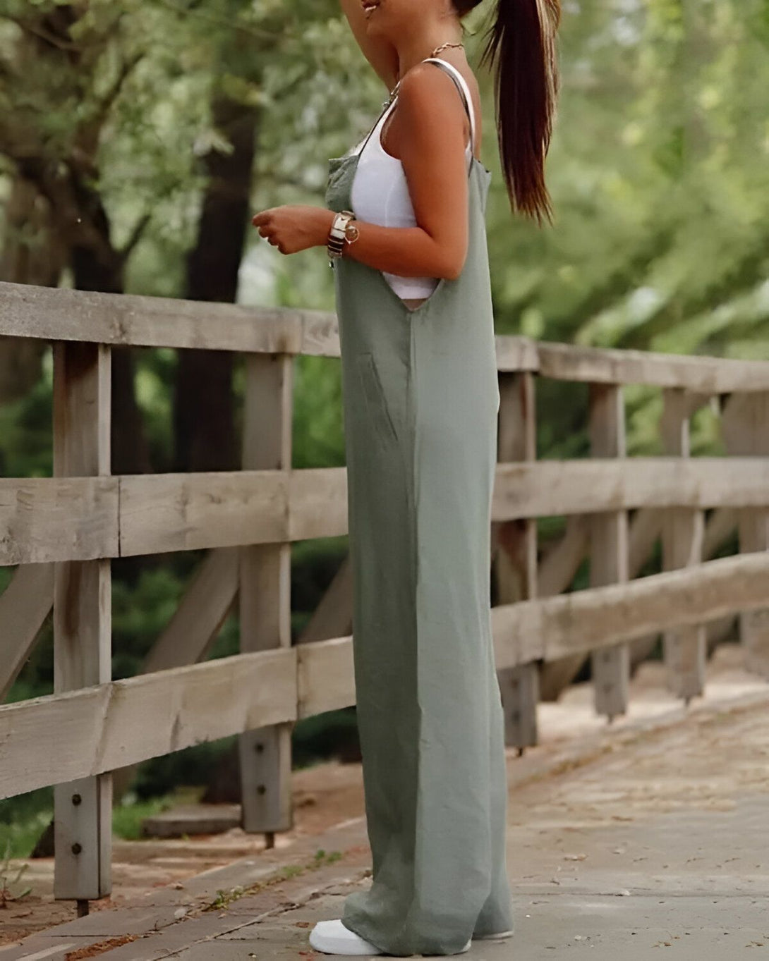 Une femme portant une combinaison-pantalon verte est debout sur un pont en bois. Elle est vue de dos et porte un sac à main marron.