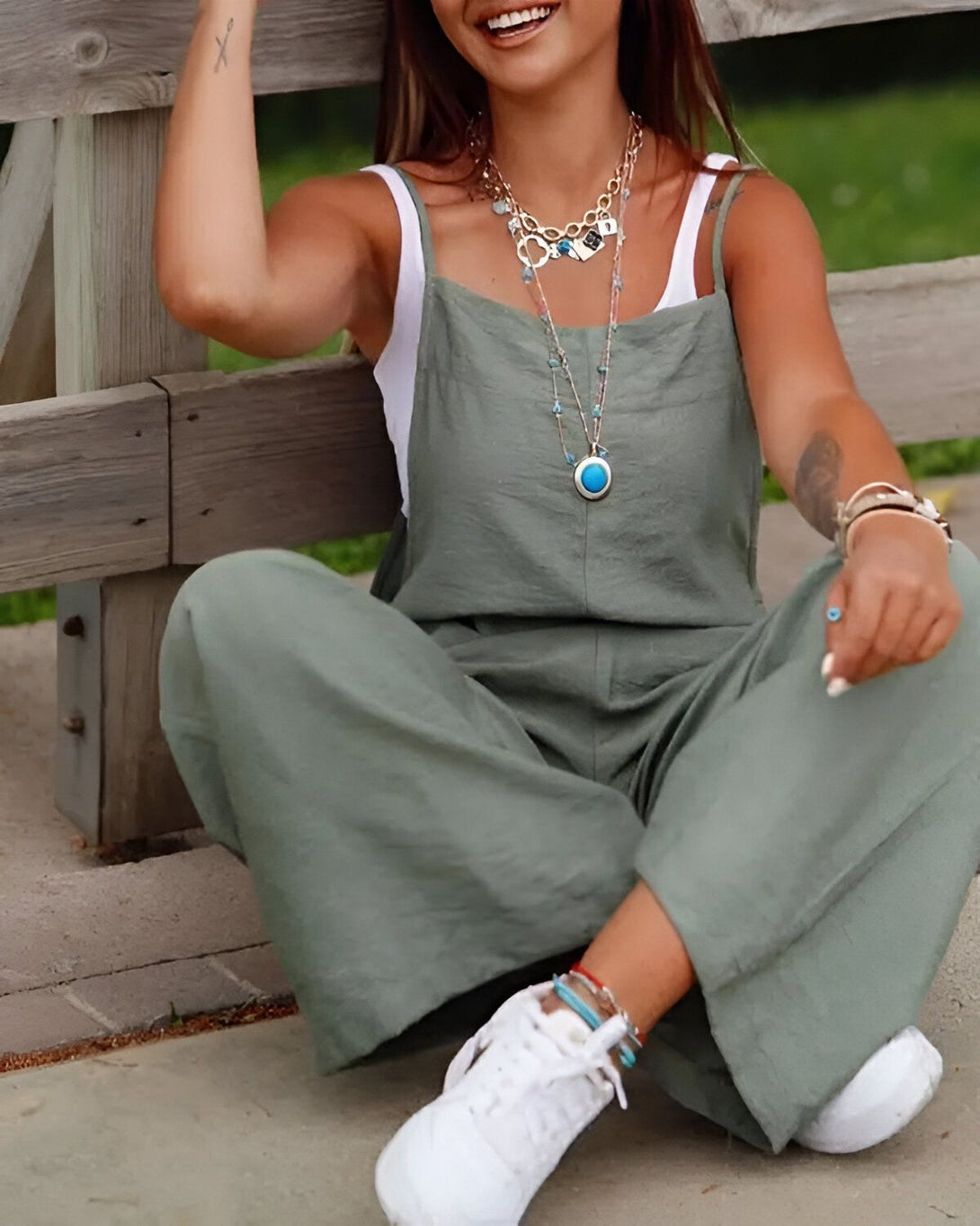 Une jeune femme est assise sur un banc en bois. Elle porte une combinaison-pantalon vert kaki, un débardeur blanc et des baskets blanches. Elle a les cheveux longs et bruns, et elle est souriante.