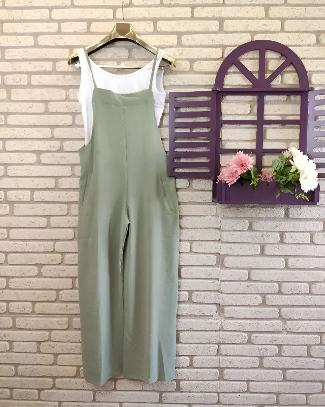Une combinaison pantalon verte est accrochée à un cintre devant un mur de briques blanches. A côté, se trouve une fenêtre en bois peinte en violet avec des fleurs artificielles roses à l'intérieur.