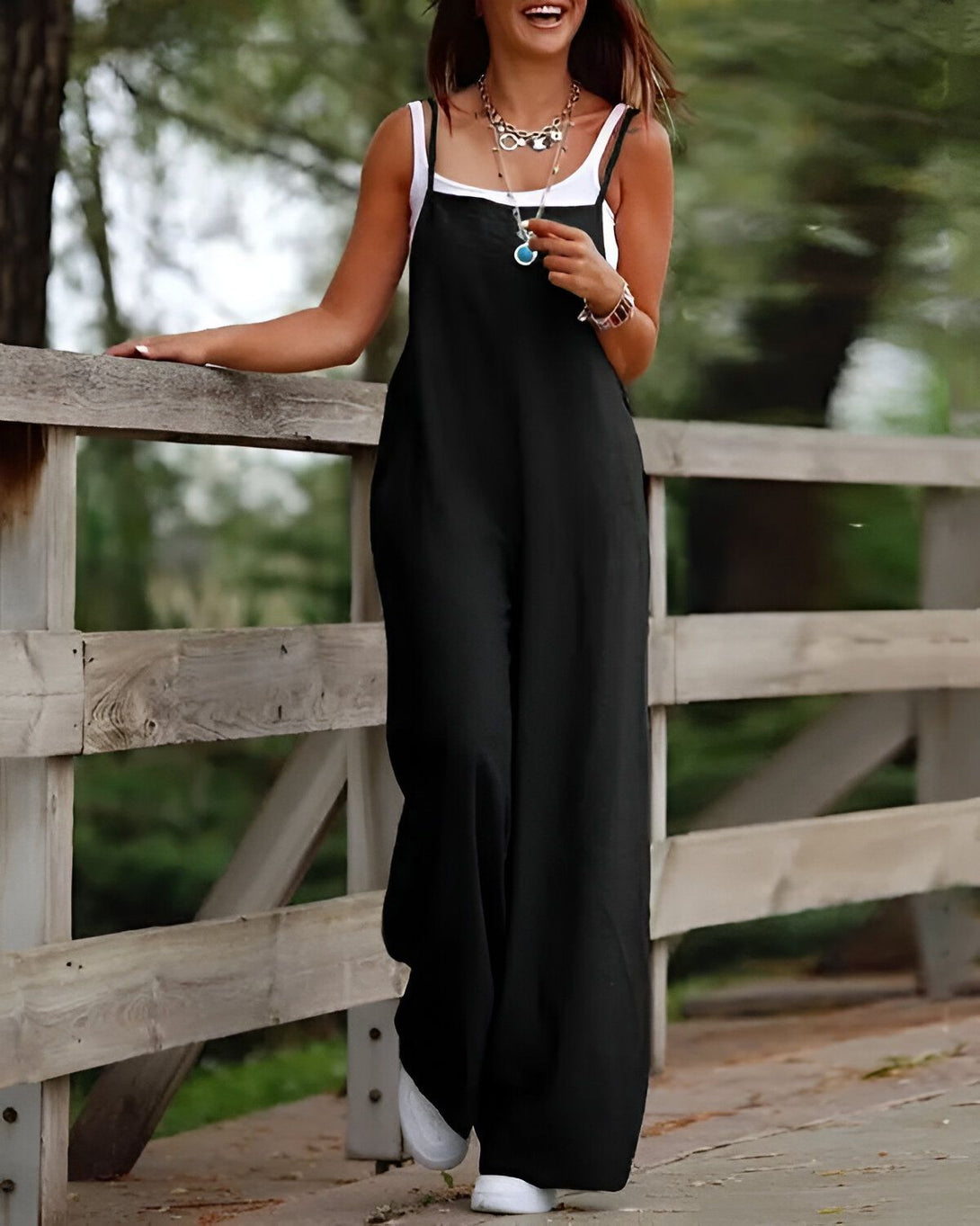 Une femme porte une combinaison noire avec des bretelles fines. Elle est souriante et se tient debout sur un pont en bois.
